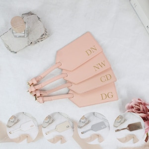 Könnte beinhalten: Vier rosafarbene Leder-Gepäckanhänger mit goldenen Buchstaben. Die Anhänger sind übereinander gestapelt und die Buchstaben auf jedem Anhänger sind: DN, NW, CD und DG.