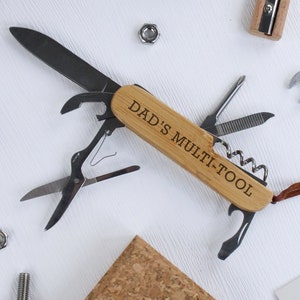 Puede incluir: Una multiherramienta con mango de madera, grabada con "DAD'S MULTI-TOOL", presenta una cuchilla, tijeras y otras herramientas. Se adjunta una correa de cuero marrón. Otras herramientas y hardware están dispersos.