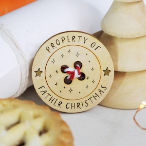 以下が含まれることがあります： 「Property of Father Christmas」という文字が書かれた木製のボタンで、赤と白の紐が穴に通されています。ボタンは白い背景で囲まれています。