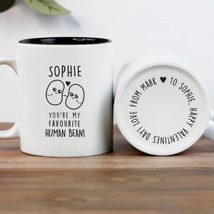 Puede incluir: Taza de cerámica blanca con borde y asa negros. La taza tiene un diseño negro de dos frijoles de dibujos animados con el texto "Sophie, ¡Eres mi frijol humano favorito!" en la parte delantera. La parte posterior de la taza tiene el texto "¡Feliz Día de San Valentín! Amor de Mark a Sophie" en un círculo.