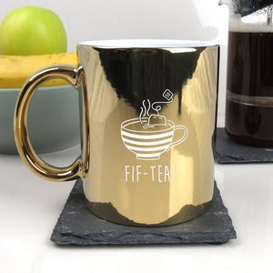 May include: Gold metallic mug with a white outline of a teacup with steam and a tea bag. The mug says "FIF-TEA!" in white lettering.