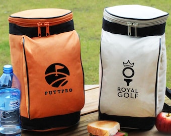 Puede incluir: Dos bolsas térmicas con temática de golf, una naranja con "PUTTPRO" y un logotipo de bandera de golf, la otra blanca con "ROYAL GOLF" y un logotipo de corona. Una botella de agua, un sándwich y una manzana sobre una superficie de madera.