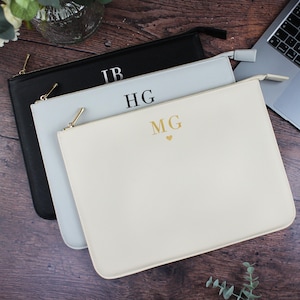 Puede incluir: Tres estuches personalizados con cremallera en negro, gris claro y crema. Cada estuche presenta iniciales doradas y una cremallera. Los estuches están colocados sobre una superficie de madera, con un portátil visible en el fondo.