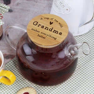 Op de afbeelding: Een glazen theepot met een houten deksel waarop staat "A cup of tea with Grandma makes everything better". Het deksel heeft een hartje. De theepot is gevuld met donkerrode thee.