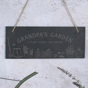 May include: Black slate garden sign with white text that reads "Grandpa's Garden, A Place Where Love Grows". The sign features a variety of gardening-themed illustrations, including a bee, boots, plants, a watering can, and a snail.