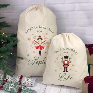 May include: Two cream-colored Christmas gift sacks with drawstring closures. One sack features a ballerina design, the other a nutcracker. Both sacks have the text "Special Delivery For" and a name in green script.