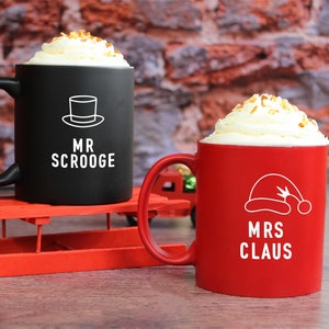 May include: Two ceramic mugs with festive designs. The black mug has a top hat and the text "MR. SCROOGE". The red mug has a Santa hat and the text "MRS. CLAUS".