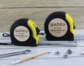 Puede incluir: Dos cintas métricas amarillas y negras con inserciones de madera. Las cintas métricas tienen el texto "Daddy No One Measures Up To You".