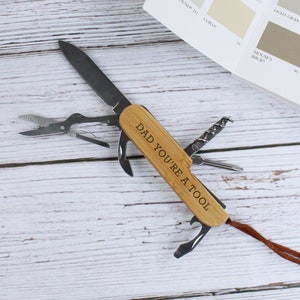 Puede incluir: Una multiherramienta con mango de madera, con varios implementos como un cuchillo y tijeras. El mango está grabado con el texto "DAD YOU'RE A TOOL". Una tarjeta de muestra de color está en el fondo.