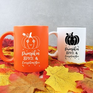 May include: Two ceramic mugs with fall-themed designs. One is orange with a white pumpkin graphic and text, the other is white with a black pumpkin graphic and text. The text on both mugs reads "Pumpkin Spice & Everything Nice."