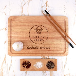 May include: A wooden cutting board with a logo that says "Chui's Chews" and the social media handle "@chuis_chews". The board is accompanied by a small wooden tray with three compartments containing red pepper flakes, salt, and black peppercorns.