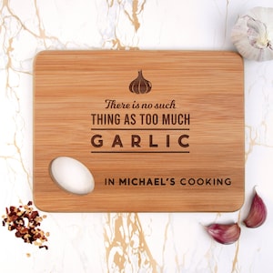 Pode incluir: Uma tábua de cortar de madeira com um desenho de alho e o texto "Não existe tal coisa como muito alho na cozinha do Michael".