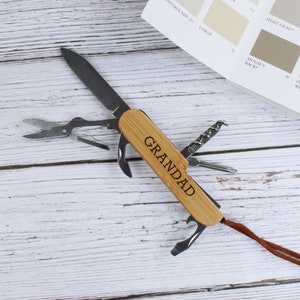 May include: A multi-tool with a wooden handle, featuring various implements like a knife, scissors, and bottle opener. The handle is engraved with the word "GRANDAD". A color swatch card is in the background.