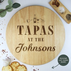 May include: A round wooden lazy Susan with the text "Tapas at the Johnsons" engraved on it. The lazy Susan is decorated with small engraved images of a wine bottle, a wine glass, and olives.