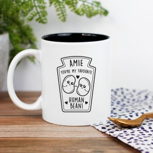 Puede incluir: Taza de cerámica blanca con borde negro. La taza tiene un gráfico en blanco y negro de dos frijoles de dibujos animados con el texto "Amie You're my favourite Human Bean!"