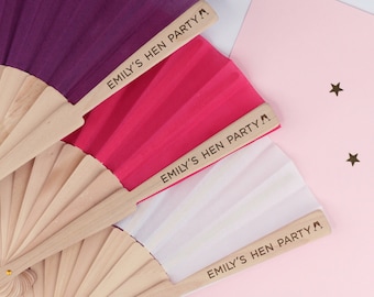 May include: Three hand fans with wooden handles and colorful fabric. The fabric is purple, pink, and white. Each fan has the text "Emily's Hen Party" printed on the handle.