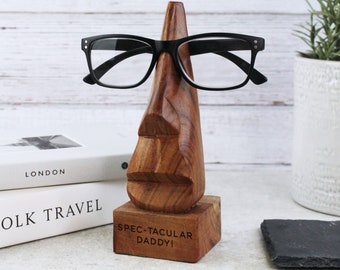 May include: Wooden eyeglasses stand shaped like a face with a nose, mouth, and eyes. The stand is engraved with the text "Spec-tacular Daddy!"