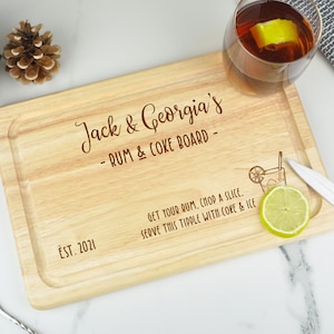 May include: A wooden serving board with the text "Jack & Georgia's - RUM & COKE BOARD - EST. 2021" engraved on it. The board also has a small illustration of a cocktail glass with a straw and a lime wedge.