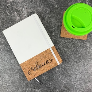 Pode incluir: Um caderno branco e de cortiça com o nome "Rebecca" escrito em itálico na capa de cortiça. O caderno está sobre uma superfície cinza ao lado de uma xícara verde com tampa.