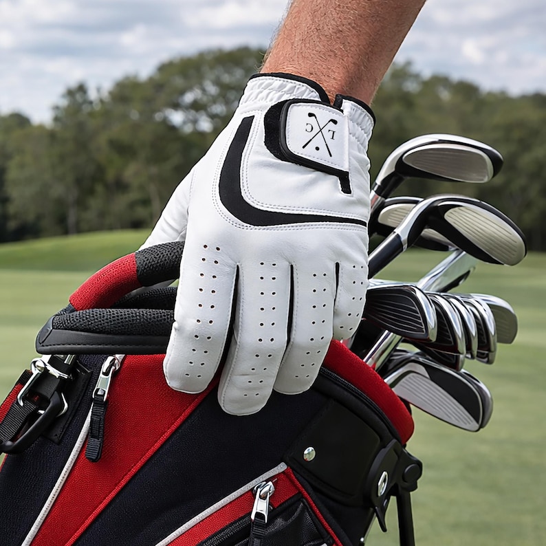 K&ouml;nnte beinhalten: Wei&szlig;er Golfhandschuh mit schwarzen Akzenten und perforiertem Design. Der Handschuh wird &uuml;ber einer roten und schwarzen Golftasche mit Golfschl&auml;gern gehalten. Der Handschuh hat ein kleines schwarzes Patch mit den Initialen "LC" und einem gekreuzten Golfschl&auml;ger-Design.