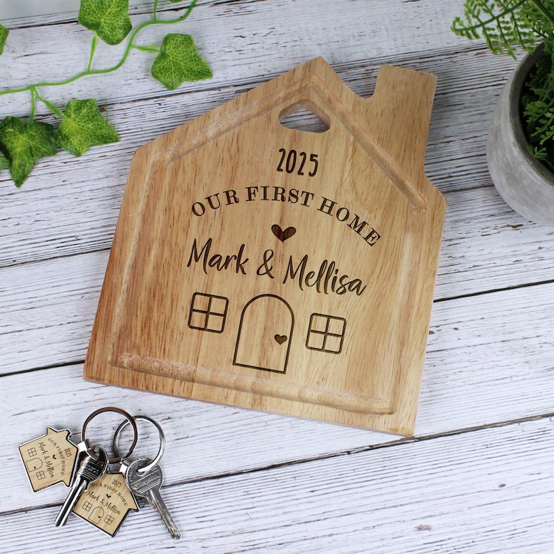 Op de afbeelding: Een houten snijplank in de vorm van een huis met de tekst "2025 Our First Home Mark & Melissa" erop gegraveerd. De plank heeft een klein hartvormig uitsparing en twee ramen.