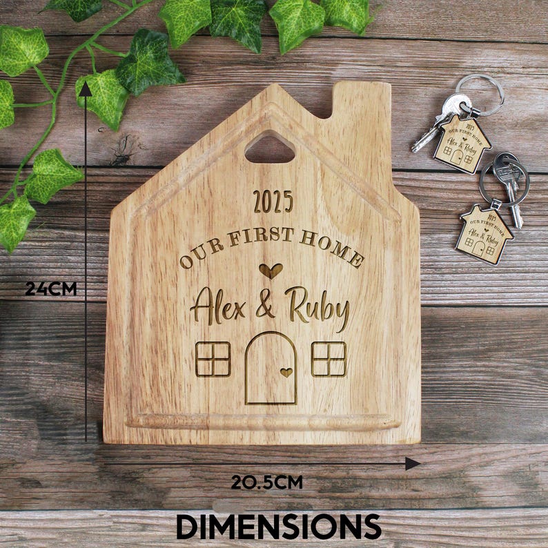 Op de afbeelding: Een houten snijplank in de vorm van een huis met de tekst "2025 Our First Home Alex & Ruby" erop gegraveerd. De plank is 20,5 cm bij 24 cm.