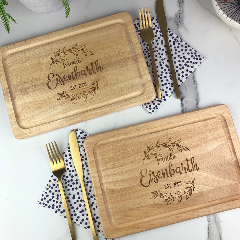 May include: Two wooden cutting boards with a decorative engraved design. The design features a wreath of leaves with the text "Familie Eisenbarth EST. 2009".