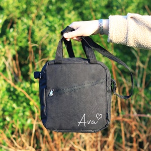 K&ouml;nnte beinhalten: Schwarze Canvas-Schultertasche mit Rei&szlig;verschlussfach und dem Namen "Ava" in wei&szlig; mit einem Herzsymbol.