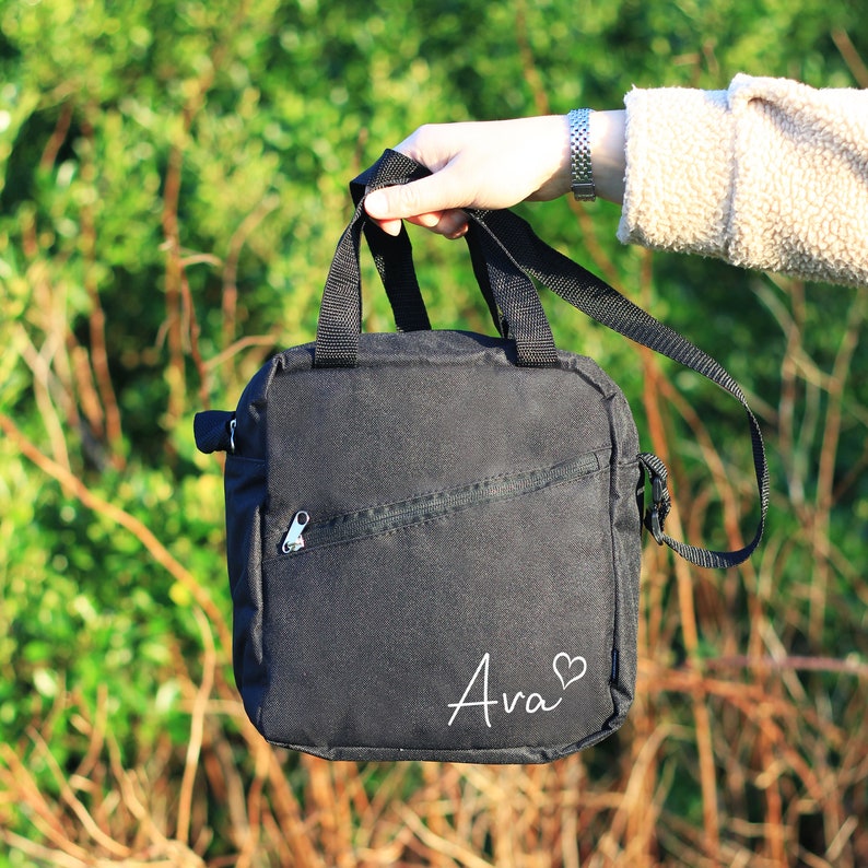 K&ouml;nnte beinhalten: Schwarze Canvas-Schultertasche mit Rei&szlig;verschlussfach und dem Namen "Ava" in wei&szlig; mit einem Herzsymbol.