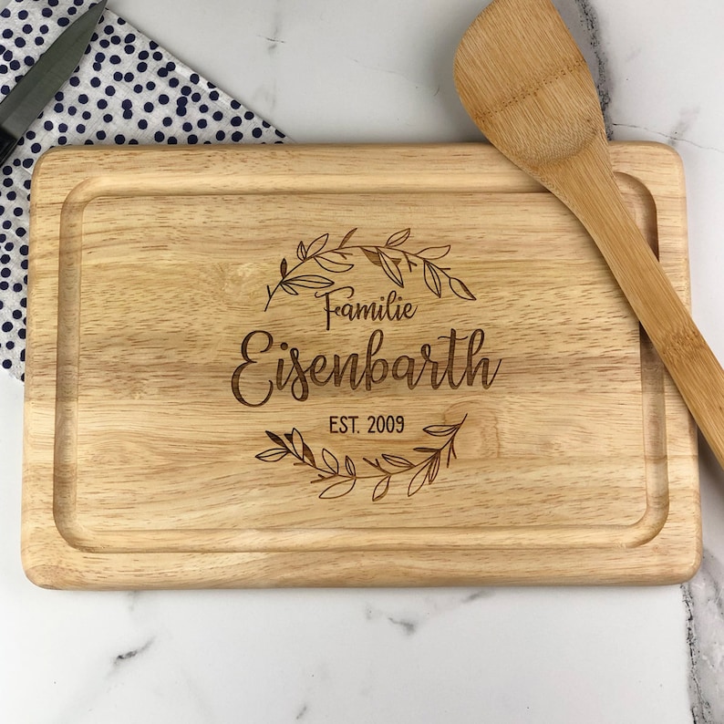 May include: A wooden cutting board with a personalized inscription that reads "Familie Eisenbarth EST. 2009" in a decorative font with a wreath of leaves around the text.