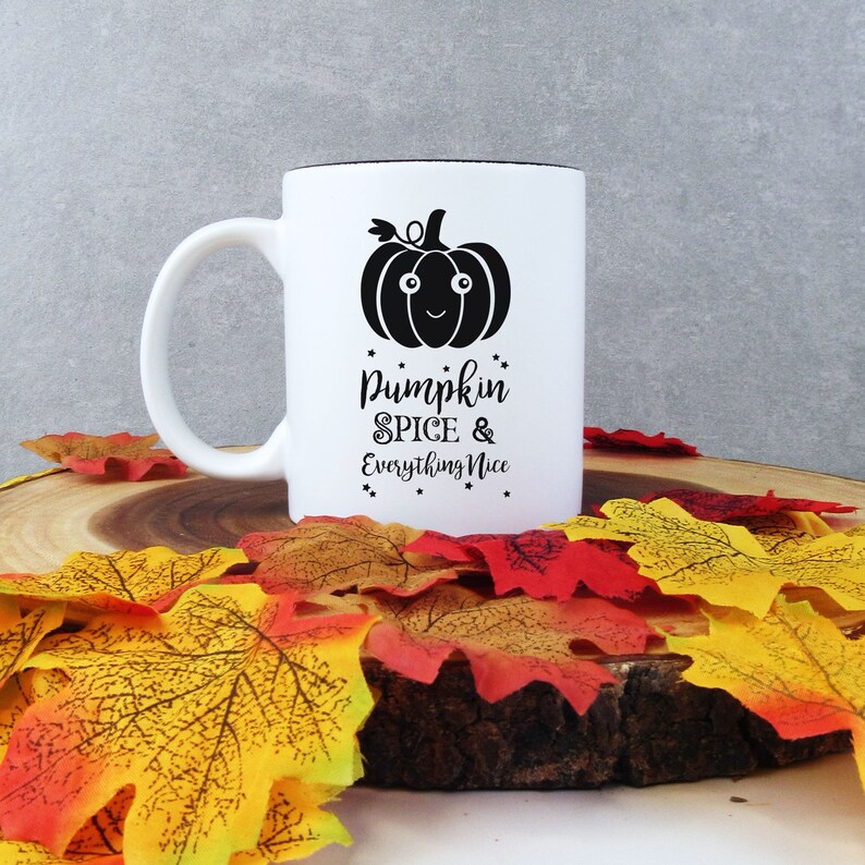 May include: White ceramic mug with a black interior and handle, featuring a black pumpkin graphic and the text "Pumpkin Spice & Everything Nice." The mug is surrounded by colorful autumn leaves, creating a seasonal theme.