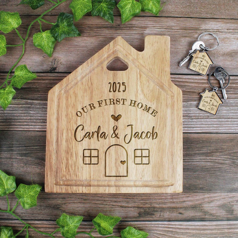 Op de afbeelding: Een houten snijplank in de vorm van een huis met de tekst "2025 Our First Home Carla & Jacob" erop gegraveerd. De plank heeft een klein hartvormig uitsparing en twee ramen aan elke kant.