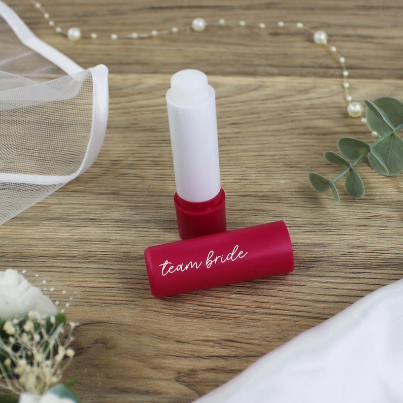 K&ouml;nnte beinhalten: Zwei rote Lippenbalsam-Tuben, eine mit wei&szlig;em Deckel und die andere mit dem aufgedruckten Text "team bride".