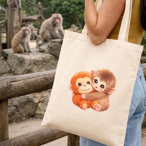 Pode incluir: Uma sacola bege com uma ilustração de desenho animado de dois macacos abraçados. Os macacos são marrons e laranja com rostos sorridentes. No fundo, há macacos reais sentados em rochas.