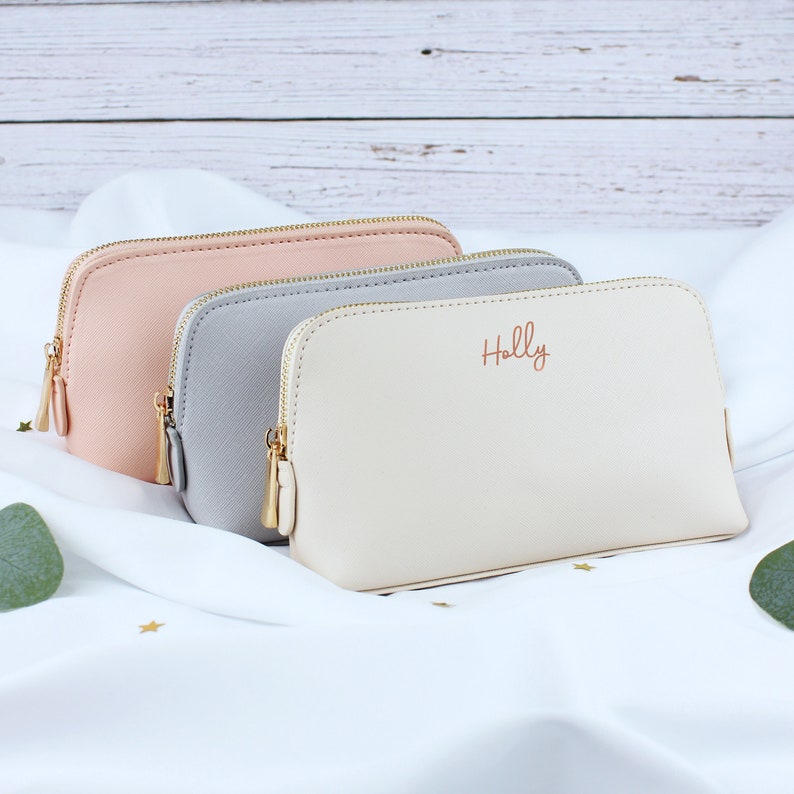 May include: Three zippered pouches in different colors: pink, gray, and cream. The cream pouch has the name "Holly" written in gold lettering.