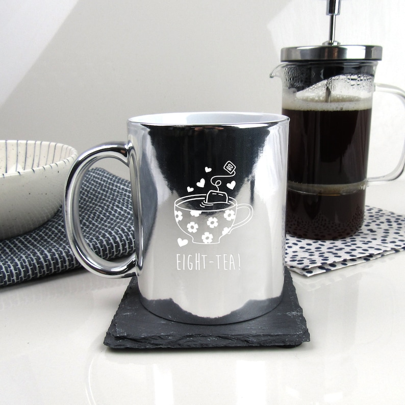 May include: A silver metallic mug with a white design of a teacup with a teabag and hearts. The text "EIGHT-TEA!" is printed below the teacup.