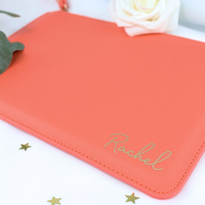 May include: A coral-coloured clutch bag with a gold zipper and the name "Rachel" written in gold script.