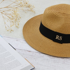 Peut inclure: Un chapeau de paille avec un bandeau noir et les initiales dor&eacute;es "RS" sur le bandeau. Le chapeau est pos&eacute; sur une surface en bois blanc.