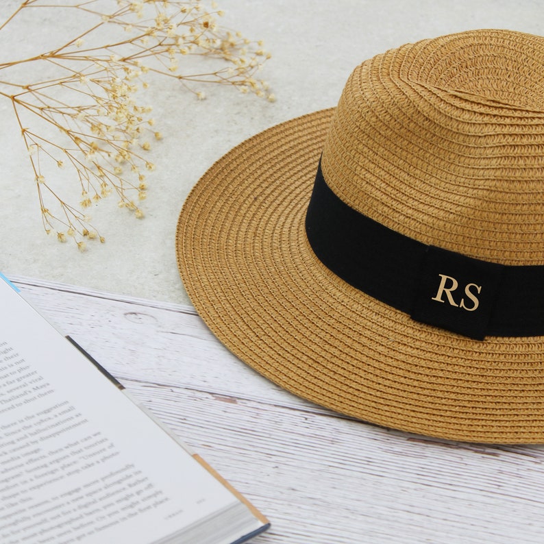 Peut inclure: Un chapeau de paille avec un bandeau noir et les initiales dor&eacute;es "RS" sur le bandeau. Le chapeau est pos&eacute; sur une surface en bois blanc.