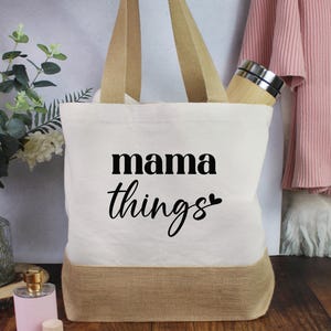 Peut inclure: Un sac fourre-tout en toile crème avec un fond et des poignées en toile de jute naturelle. Le sac porte les mots "mama things" en écriture noire et un cœur. Un vase de fleurs blanches, un flacon de parfum et un pull rose sont en arrière-plan.