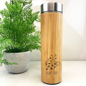 May include: A bamboo travel mug with a stainless steel lid. The mug has a laser engraved design of a teacup with a tea bag and the text "Eight-Tea!"
