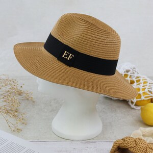 Peut inclure: Un chapeau de paille brun avec un ruban noir et les initiales dor&eacute;es "EF" sur le devant. Le chapeau est pos&eacute; sur une t&ecirc;te de mannequin blanche.