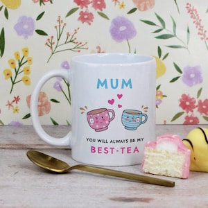 May include: White ceramic mug with the word "MUM" in blue, and the phrase "YOU WILL ALWAYS BE MY BEST-TEA". The mug features illustrations of two teacups and hearts. A gold spoon and pastries are in the foreground.