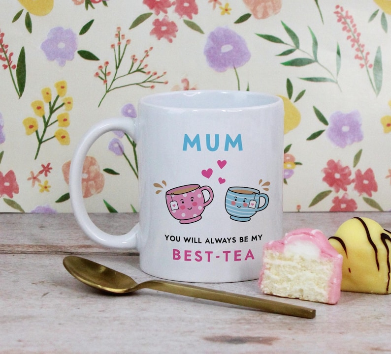 May include: White ceramic mug with the word "MUM" in blue, and the phrase "YOU WILL ALWAYS BE MY BEST-TEA". The mug features illustrations of two teacups and hearts. A gold spoon and pastries are in the foreground.