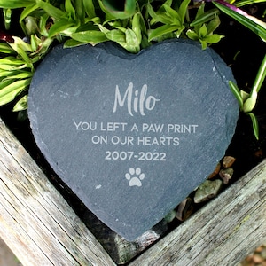 Puede incluir: Una piedra conmemorativa de pizarra en forma de corazón con el nombre "Milo" grabado en ella. La inscripción dice "You left a paw print on our hearts 2007-2022" e incluye un símbolo de huella de pata.