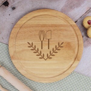 May include: Round wooden cutting board with a groove around the edge. The board features a brown illustration of a whisk, spatula, and a laurel wreath. The board is light brown and sits on a green and white checkered cloth.