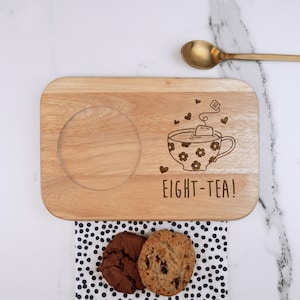 May include: Wooden serving tray with a cutout for a mug, a teacup with a teabag and hearts, and the text "EIGHT-TEA!" engraved on the surface. Two chocolate chip cookies are on a black and white polka dot napkin.