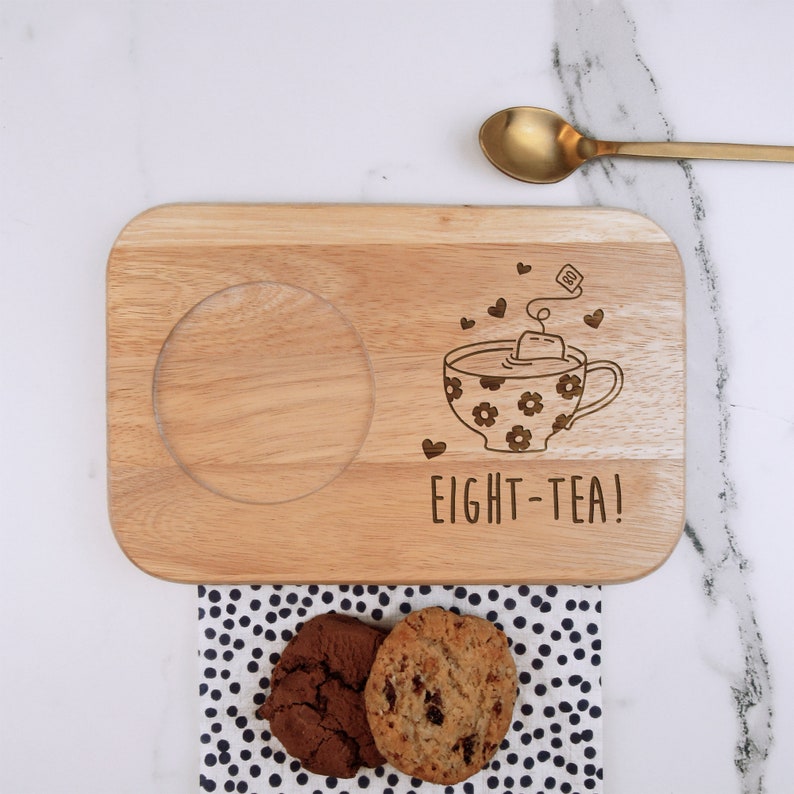 May include: Wooden serving tray with a cutout for a mug, a teacup with a teabag and hearts, and the text "EIGHT-TEA!" engraved on the surface. Two chocolate chip cookies are on a black and white polka dot napkin.