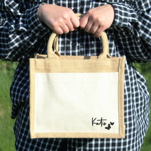 May include: A natural jute tote bag with a white canvas insert. The bag has a black script font that says "Katie" with two black butterfly silhouettes.