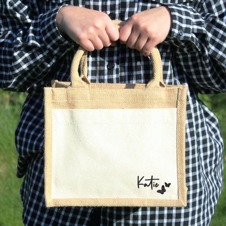 May include: A natural jute tote bag with a white canvas insert. The bag has a black script font that says "Katie" with two black butterfly silhouettes.